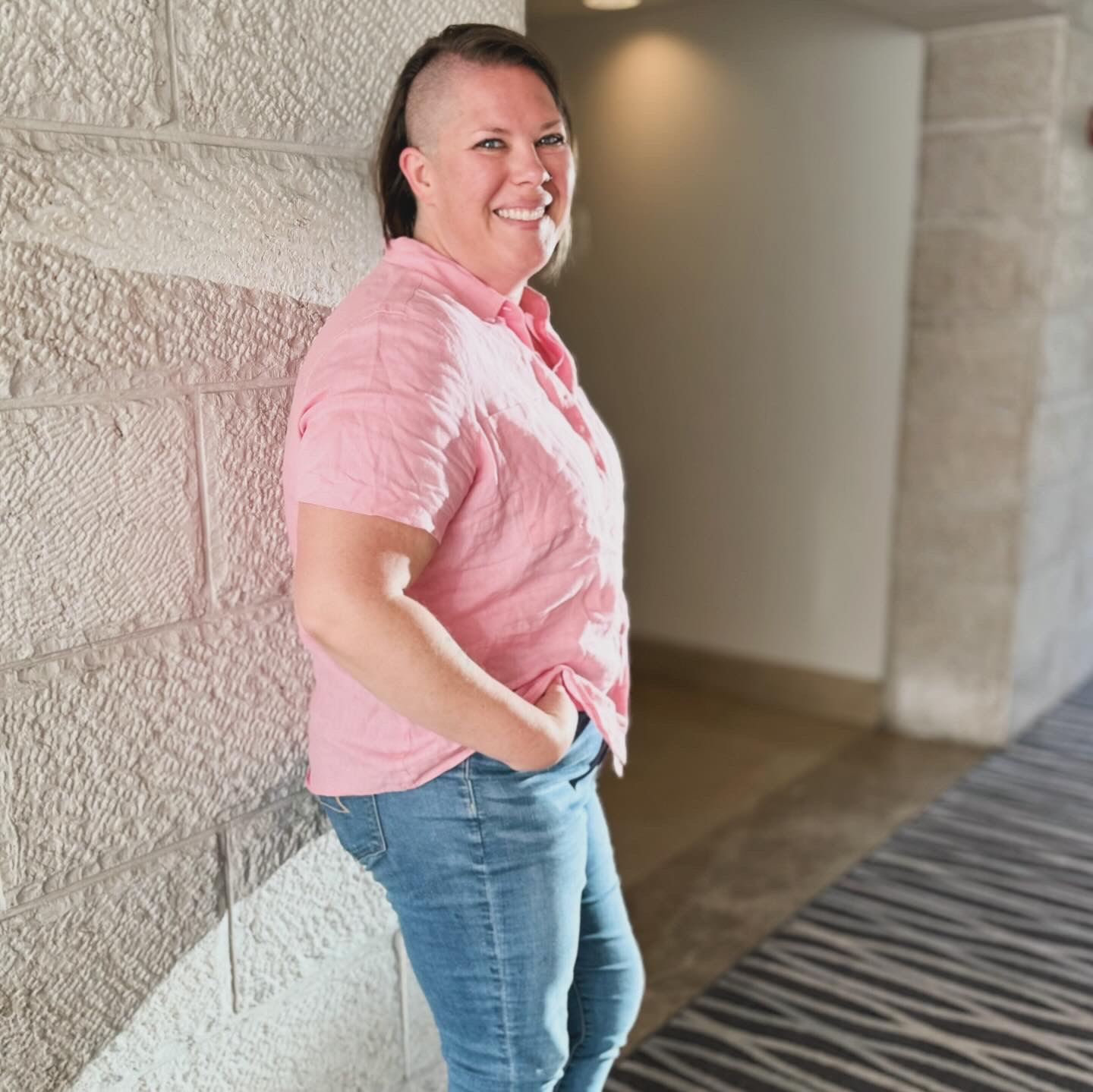 Kimberly in a pink shirt and jeans leaning against a brick wall